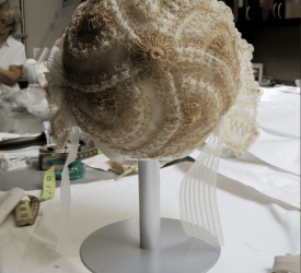 Backside of a cream lace cap embroidered with raised gold swirling motifs and scalloped edging, mounted on a display stand in a studio workspace.